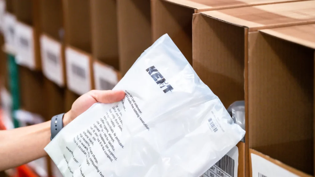 Hand holding a poly mailer shipping bag beside labeled warehouse storage bins